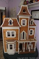 Front view of Victorian style wooden dollhouse showing bay window, multiple roof levels with shingle detailing, white window shutters, and front porch columns.