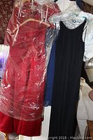 Photo showing multiple formal dresses covered in plastic, including red and black ones hung on wooden hangers.