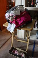 Pile of assorted women's clothing folded and stacked on a small folding wooden table. Variety of colors including bright pink, gray, white, and black. Some items with tags attached.