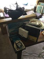 Lot includes Omega Mini Steam Zapper in box, two unopened photo albums, Hallmark mother of pearl 4x6 frame in box, and rolls of wrapping paper, arranged on a table.