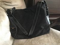 Black suede and leather shoulder bag with white stitching resting on a cushioned surface