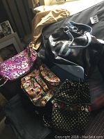 Photo showing multiple bags including black leather-look bag with blue accent handles, purple quilted floral bag, black fabric floral embroidered bag, and multicolored pattern shoulder bag.