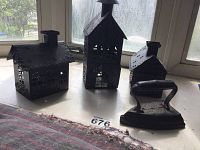 Three black cast iron tin houses of varying sizes with detailed designs and opening doors, displayed on a windowsill alongside a vintage cast iron clothes iron.