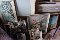 Assorted framed wall decorations stacked, showing a floral painting, additional prints, and artworks including a small piece with human figures and abstract details.