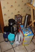 Wide view of the six assorted thermos and hot beverage cups, Black and Decker hand mixer, and boxed cookie cutters set on a tiled floor in front of patterned wallpaper.