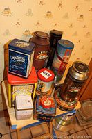 A group shot showing multiple tins of various brands and shapes including Maxwell House coffee, Baileys Irish Cream, and other assorted tins arranged on a small wooden stand.