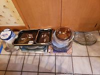 Overview photo showing glass bakeware, mixing bowls, rectangular metal trays with floral designs stacked on tiled floor.