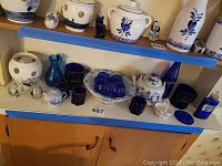Shelf with various blue glass bottles, blue glass mugs, white ceramics with blue designs including a tea kettle, decorative ceramic figurines, and a woven basket with blue glass items.