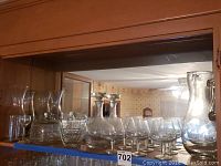 Glassware assortment including six goblet glasses, two decanters with stoppers, footed pastry dish, and large etched pitcher on a shelf.