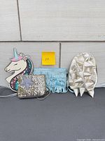 Front view of all four items: unicorn purse, fringe purse, sequin coin purse, dinosaur backpack