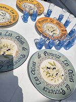 Overview of five Provence Les Olives plates and eleven Three Olives blue tall shot glasses on table