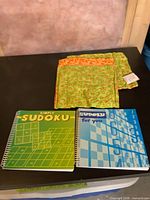 Two Sudoku books and folded stack of patterned microwave potato holders on table