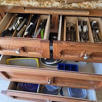 Open kitchen drawers showing knives, flatware, utensils, lower drawers with bowl, colander, lids, ice cube tray