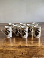 Eight ceramic mugs with holly motif and gold accents on wooden surface