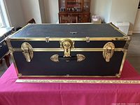 Front view of closed trunk showing black faux leather exterior, brass hardware, central lock, side latches, handle
