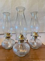 Front view of three matching clear glass oil lamps showing ribbed bases, brass burners, and chimneys