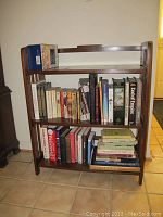 Front angled view of collapsible wooden bookshelf with three shelves filled with various books, showing condition and shelving layout.