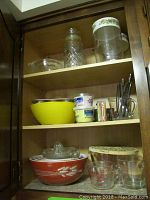 Photo showing upper and middle shelves in wooden cabinet with glass jars, plastic bowl, cupcake wrappers, Pyrex bowls and utensils.