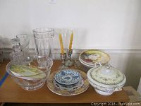 Overview of lot on wooden table showing various glass, china, crystal, and decorative items including bowls, plates, vases, decanter, and candlesticks