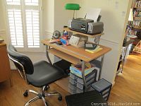 Full view of desk with metal frame, top shelf holding printer, pull out keyboard tray, lower shelves, and blue office chair with caster wheels in front