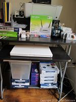Overview of items on the black desk including HP Deskjet printer, TP-Link router box, ink cartridges arranged, folders, and office supplies.