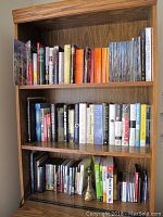 Full view of wooden bookshelf with assorted books on three shelves including history, political, fiction, business, and opera encyclopedia books plus brass bookends