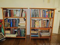 Two wooden shelves filled with assorted hardback and softback books of various sizes and colors on a hardwood floor against a white wall with power outlet and door visible.