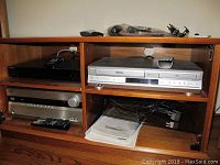 Wide shot of wooden media cabinet displaying the three electronic devices and several remotes on and inside compartments.