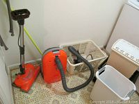 Kenmore Hepa 12 amp canister vacuum with black hose and orange-red body, vacuum attachment, plastic hamper, white waste can, and a beige waste can with floral lid design against a patterned linoleum floor.