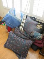 Photo showing the blue and white patchwork quilt draped over a large woven basket along with pillows displaying paisley and floral patterns. Several 65-inch white metal curtain rods are standing upright.
