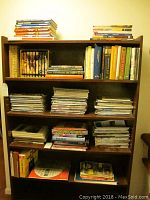 Bookshelf showing stacks of hardcover books and loose magazines on multiple shelves