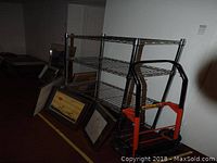 Chrome bakers rack with three wire shelves beside a black and orange convertible dolly in an indoor setting with framed pictures leaning against the wall.