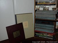 Stacked Metropolitan Museum of Art portfolios and various art history books on a shelf, showing titles related to pre-Columbian art and world history atlas.
