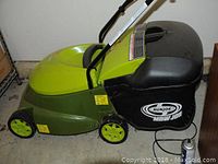 Side and partial front view of green electric lawn mower with black grass collection bag attached, shows wheels and some wear.