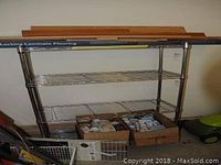Full view of the chrome bakers rack showing three wire shelves and vertical posts with wood pieces stored on top shelf and boxes underneath.