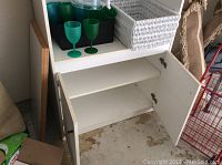 Photo of white shelving and cupboard storage cabinet partially open showing interior shelves and contents inside including plastic wine glasses, wicker basket, and drink container.