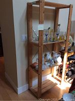 Wood shelving unit in natural unfinished wood with multiple shelves holding glassware, jugs, a small baker, and sake bottles under wall light near corner.