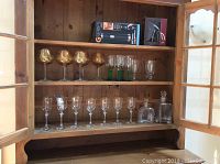 Wide view of a wooden cabinet with three shelves containing various wine and brandy glasses, a boxed G Series auto wine opener, decanter, and corkscrew.