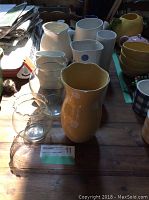 Image shows variety of ceramic and glass vases on a table with sunlight and shadows, including yellow vase in foreground, white pitchers and clear glass vases.