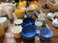 Photo showing a collection of colorful ceramic tableware including jugs, teapots, bowls and mugs arranged on a wooden table.