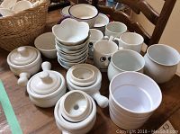 Photo showing white ceramic pieces including coffee mugs with reindeer, lidded soup bowls, lotus shaped bowls, napkins with rings and a basket of dishes on a wooden table.