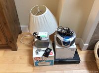 Photo shows white shade table lamp, IKEA Astrid lamp box, silver Life humidifier, Philips CD player and radio, and Goldstar VHS player all grouped on a hardwood floor.