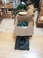 Metal Christmas tree stand, cardboard box with Christmas lights and garlands, wicker baskets behind