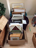 Photo showing a box and plastic bin filled with multiple picture frames in assorted sizes and finishes, including a distinctive house-shaped frame.