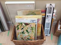 Photo showing a wicker basket filled with several original paintings and framed art pieces, different sizes, some floral themes.