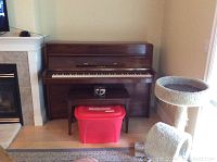 Full view of Yamaha upright piano, bench, and red Rubbermaid container with music books next to a cat tree and fireplace.
