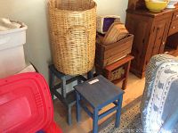 Blue painted wooden step stool and side table with visible wear next to a round natural wicker laundry hamper and wooden crate with wicker baskets.