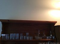 Glass jug, various clear glass tumblers and drinking glasses, and a glass cake stand arranged on a wooden shelf.