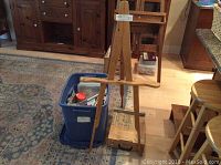 Wooden art easel standing on a hardwood floor with a blue Rubbermaid container of art supplies and a wooden painter's box on the floor nearby.