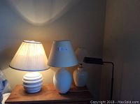 Four lamps on wooden surface against wall: brass telescopic floor lamp, two white ceramic table lamps with shades, small black shade reading lamp